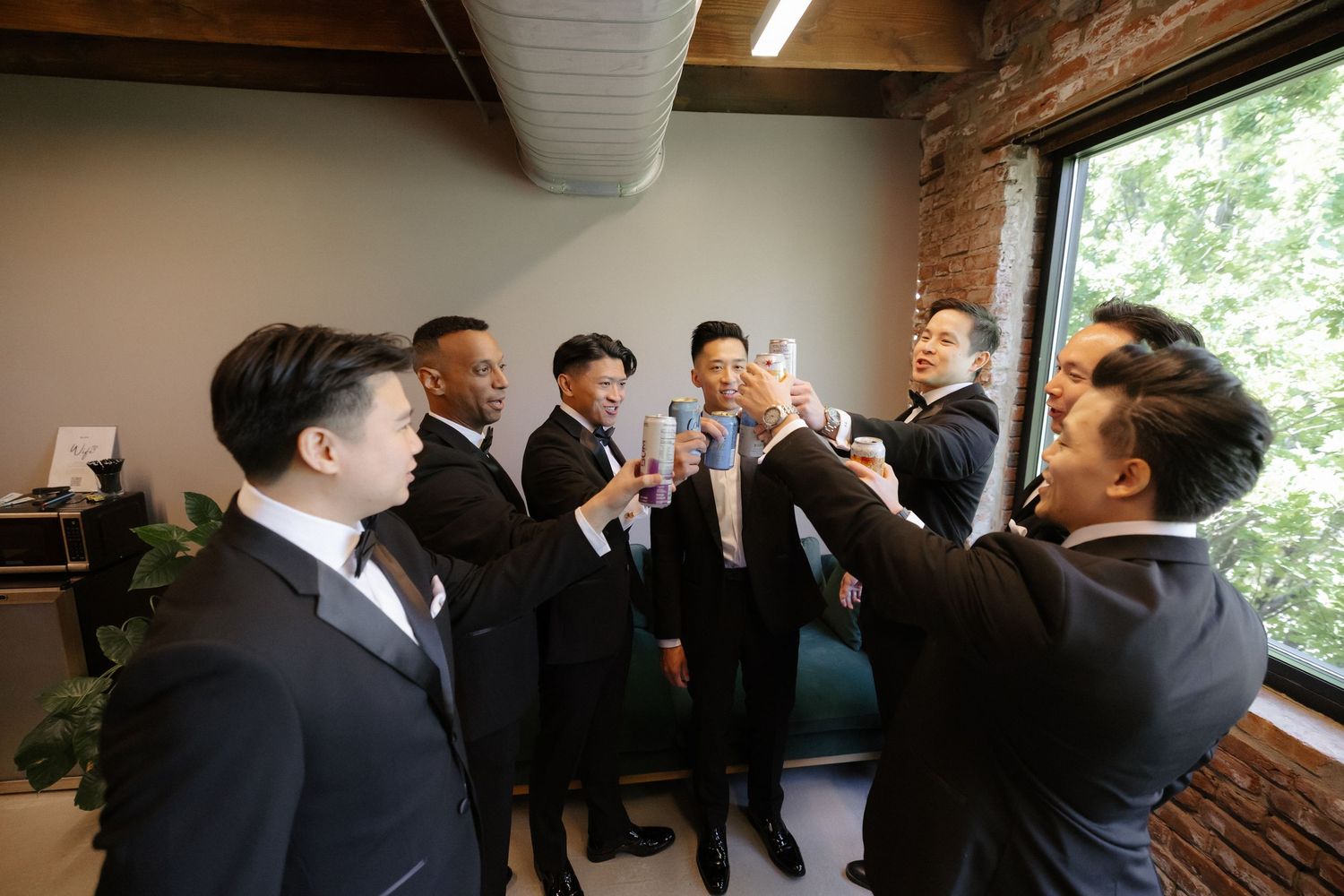 A group of groomsmen in black suits toasting drinks together in a modern room with brick walls and large windows.