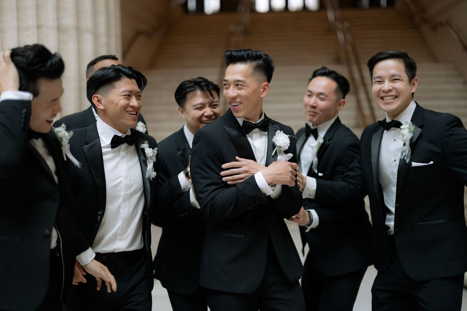 Groom laughing with groomsmen in black tuxedos on marble stairs at an elegant wedding venue.