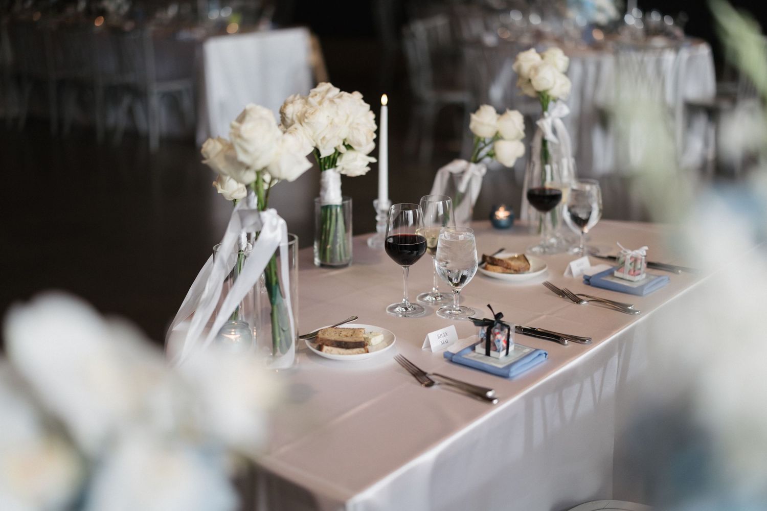 Elegant wedding reception table with white floral arrangements, candles, wine glasses, and a bride and groom cake topper.