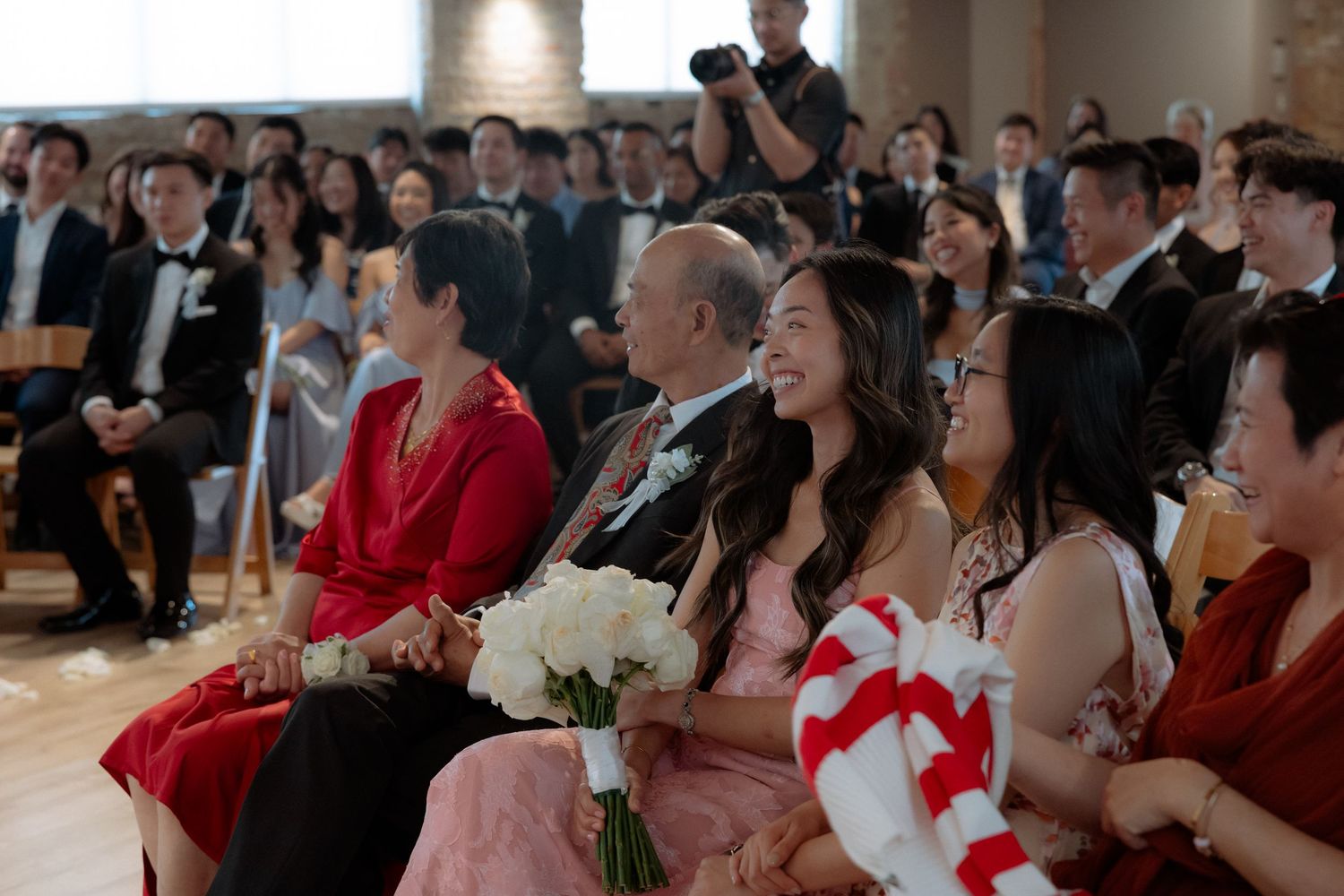 Guests smiling and watching a wedding ceremony, with a woman in pink holding white flowers seated in the front row.