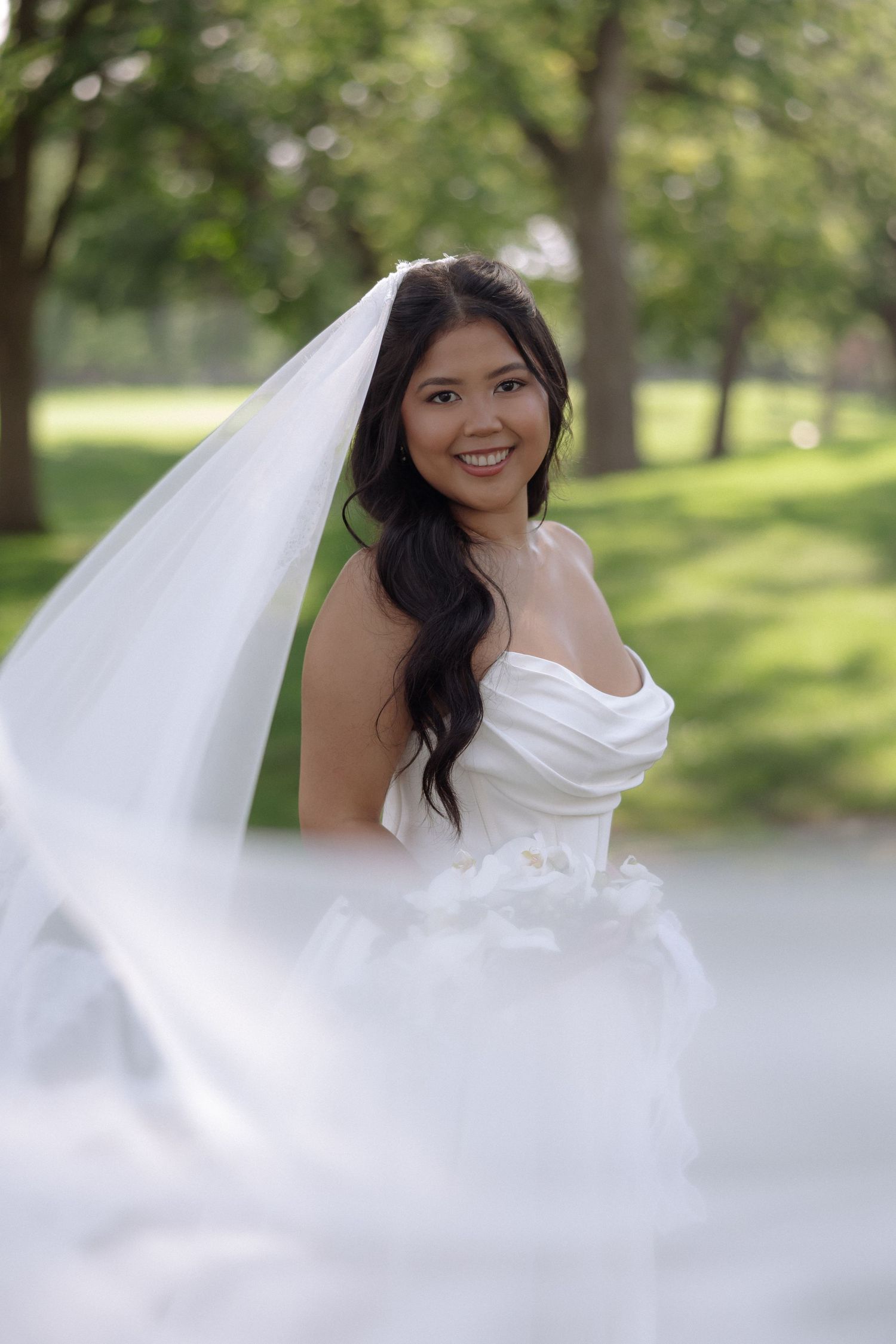 Smiling bride in strapless white wedding dress and veil posing in a lush green park setting.