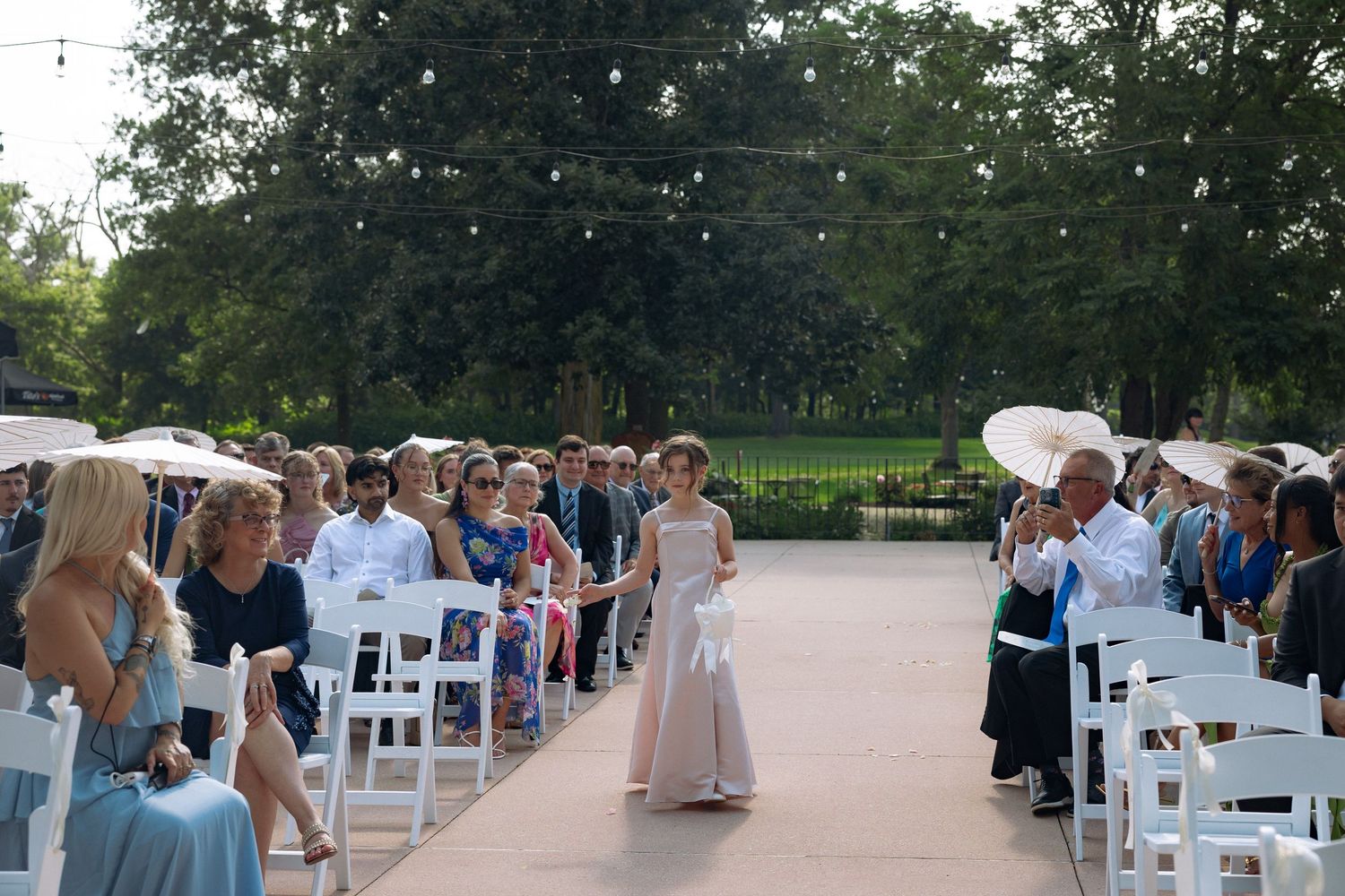 Young flower girl walks down outdoor wedding aisle in elegant gown, guests seated in white chairs under string lights.