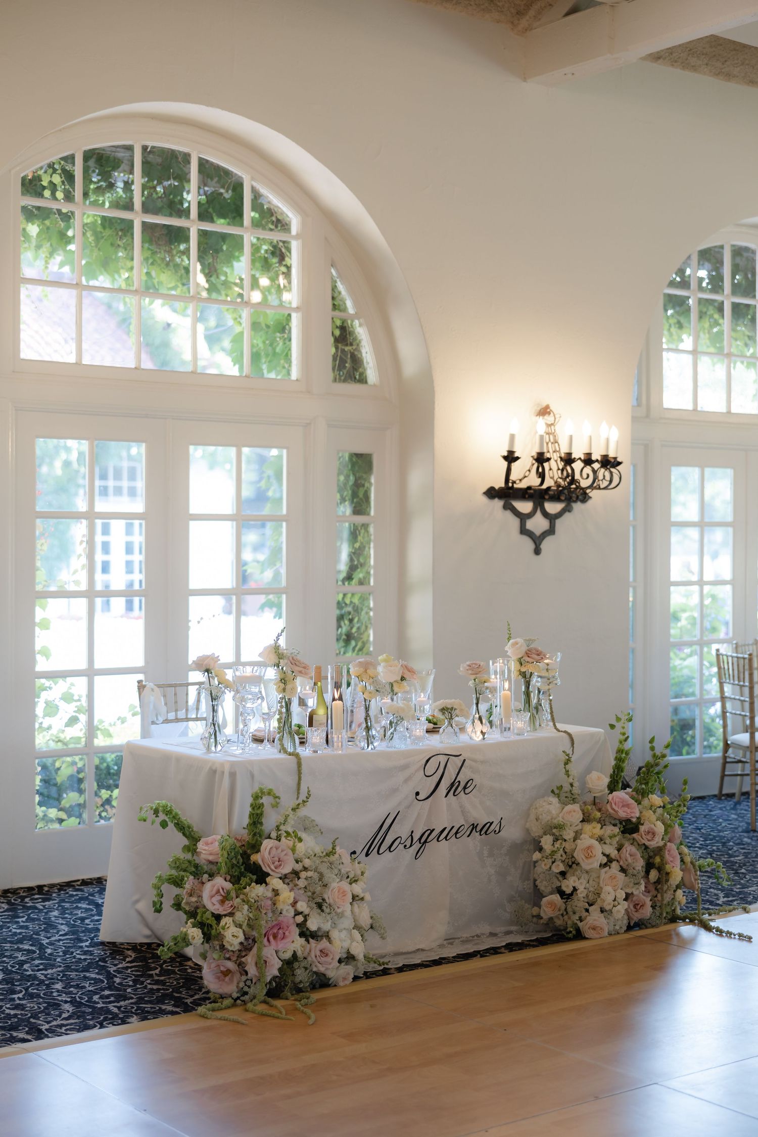 Elegant sweetheart table with floral arrangements and The Mosqueras sign in a bright white ballroom.