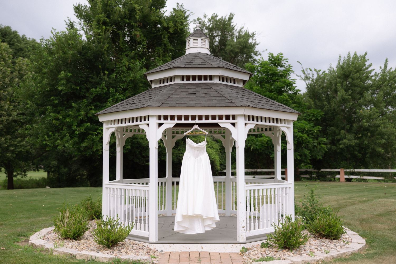 White wedding dress hanging in a charming white gazebo surrounded by lush green trees on a sunny day.