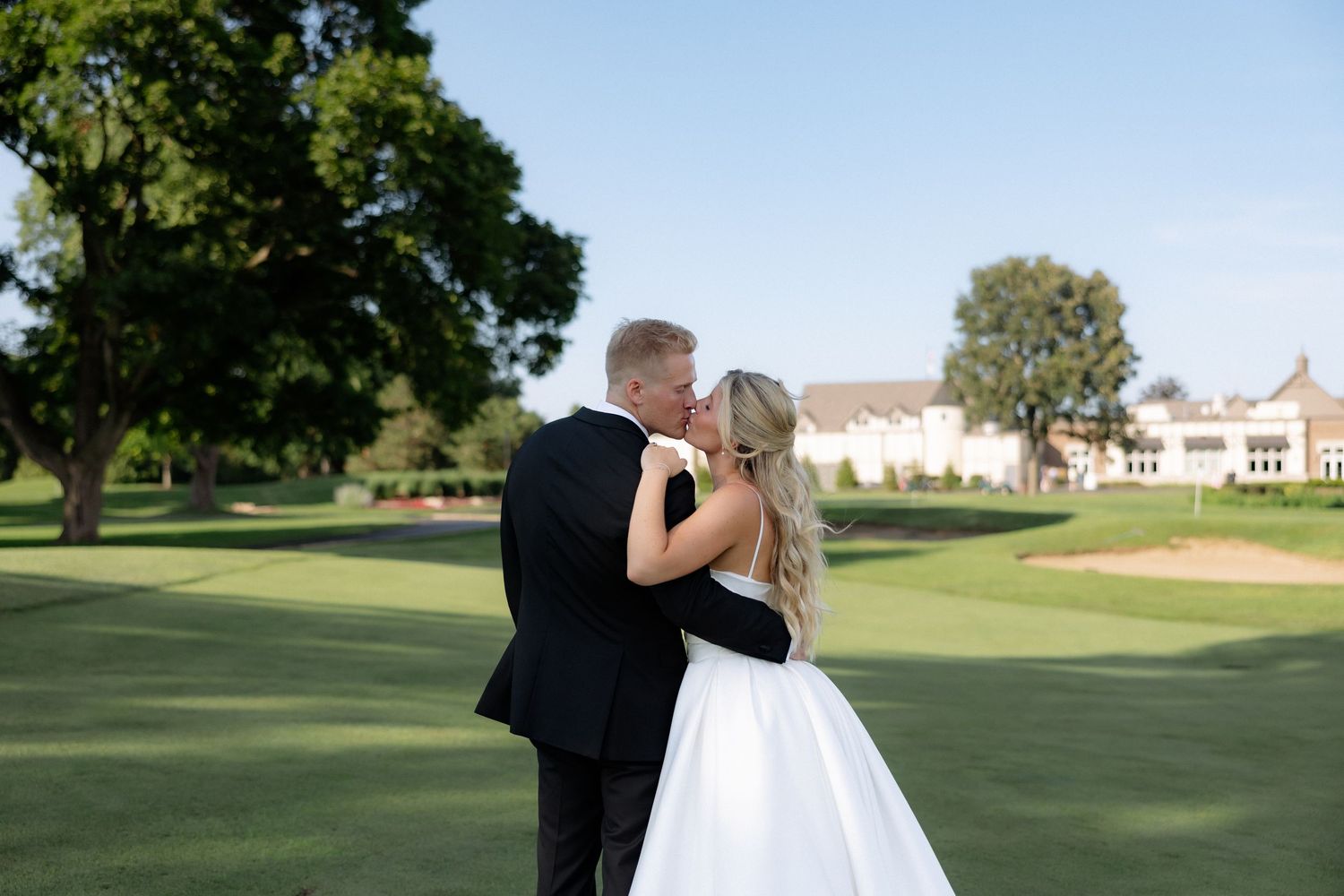 Bride and groom share a kiss on a scenic golf course with a grand clubhouse in the background.