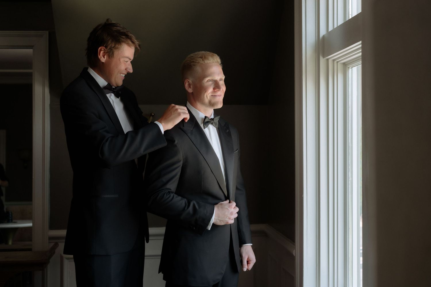 Father helping groom adjust tuxedo jacket near bright window before wedding ceremony.