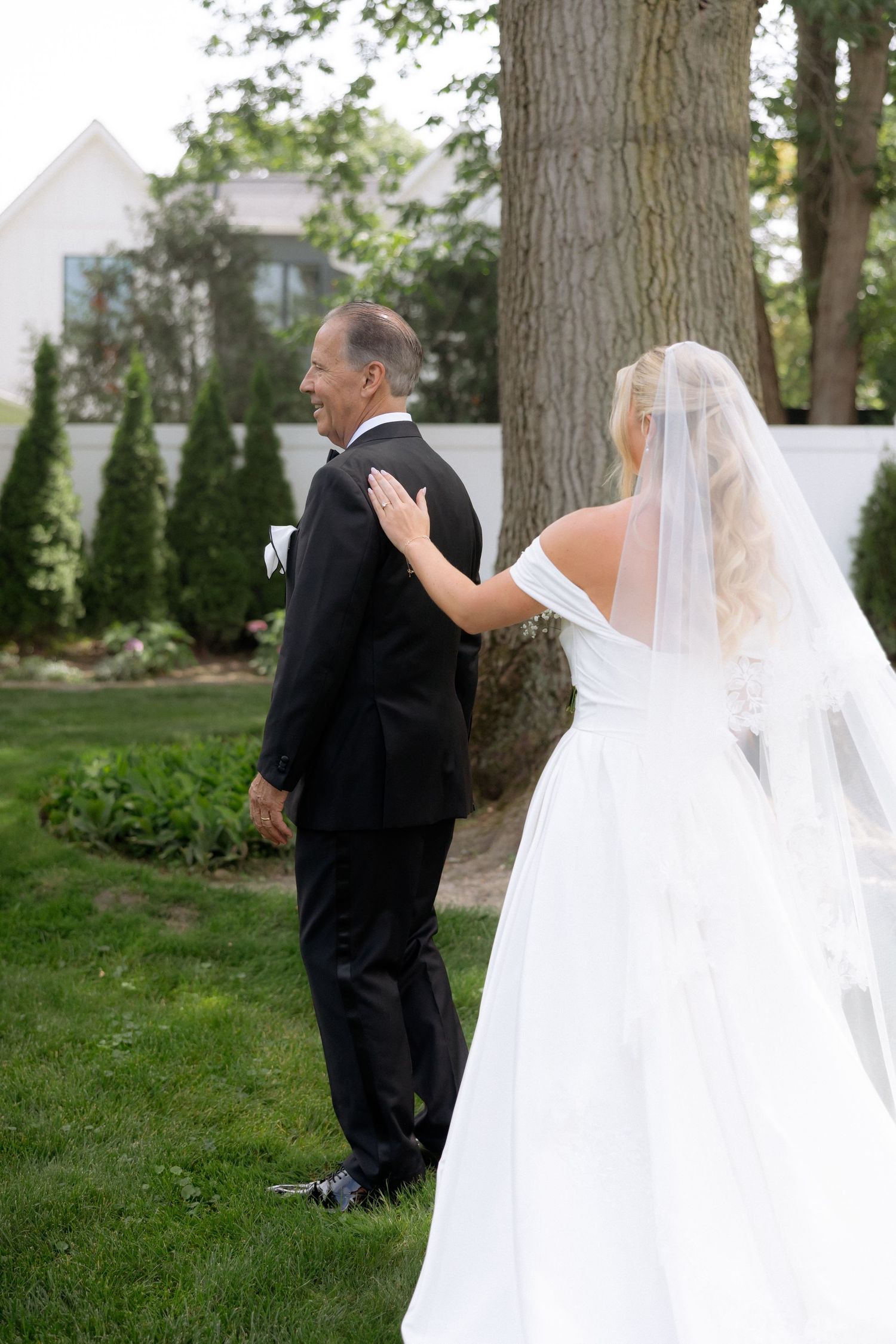 Bride in white gown with veil taps groom on shoulder for first look in lush garden setting.