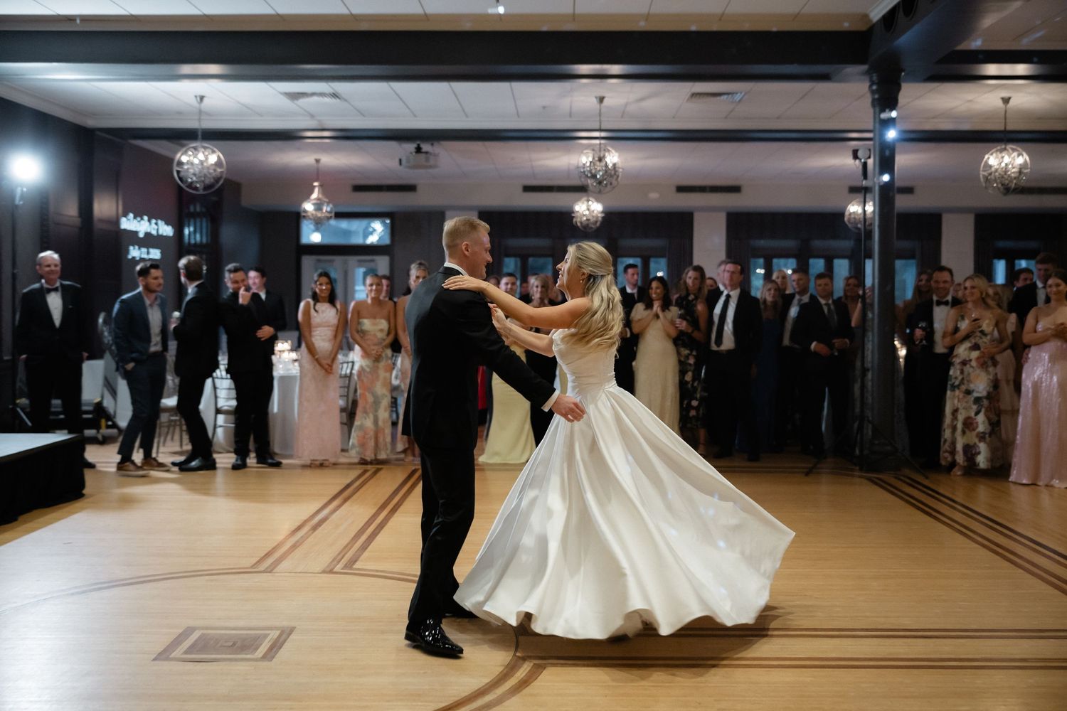 Bride and groom sharing romantic first dance on ballroom floor surrounded by wedding guests.