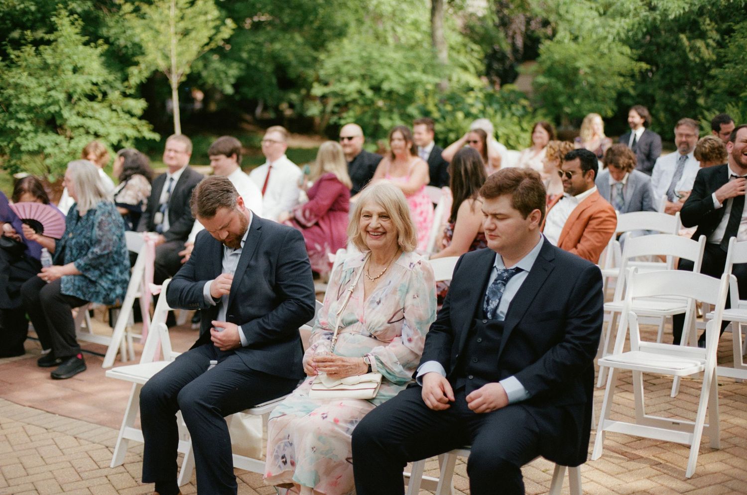 Guests seated in white chairs at an outdoor wedding ceremony surrounded by lush green trees.