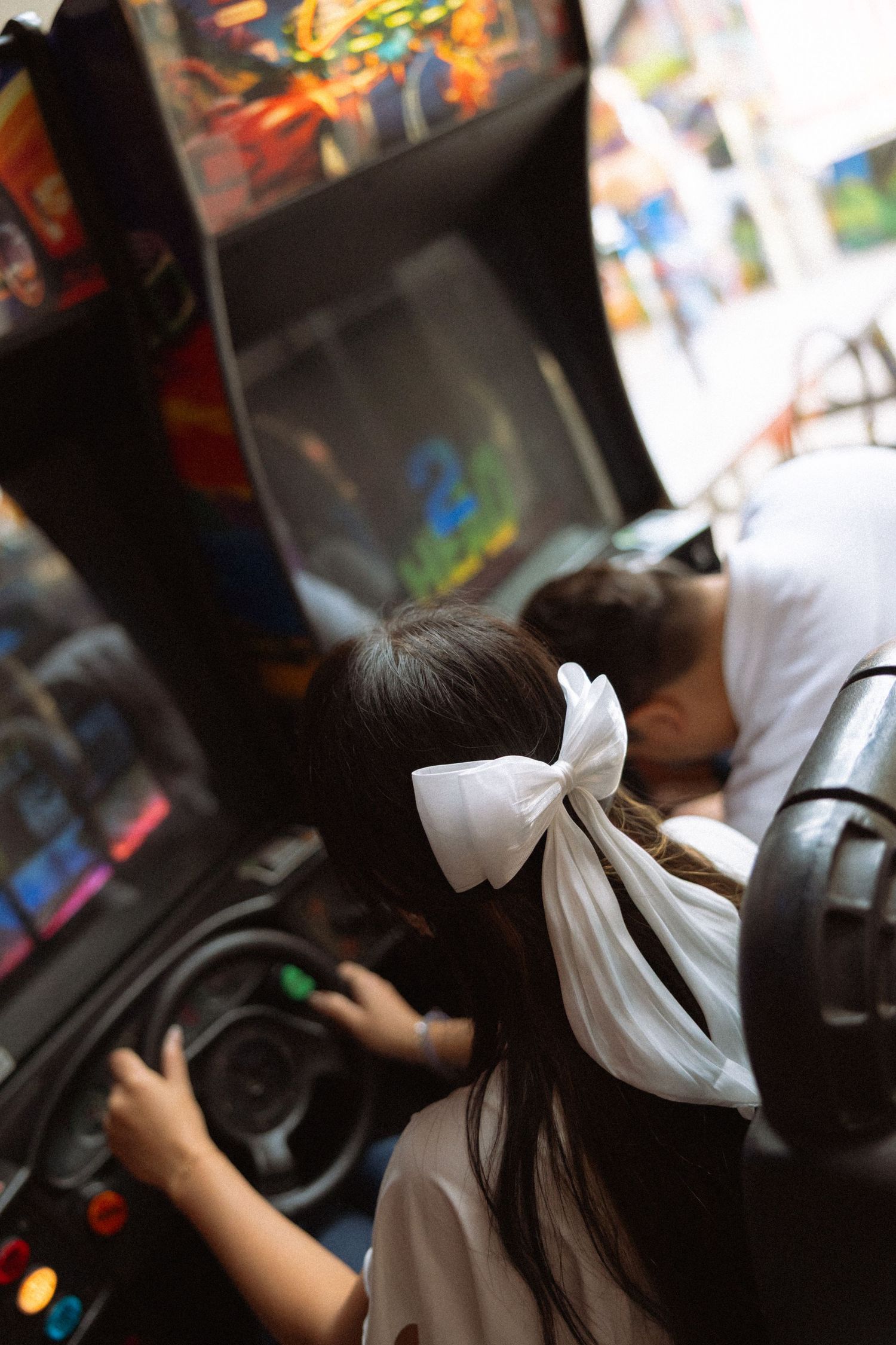 Young girl with white bow plays arcade racing game, viewed from behind in colorful game center.