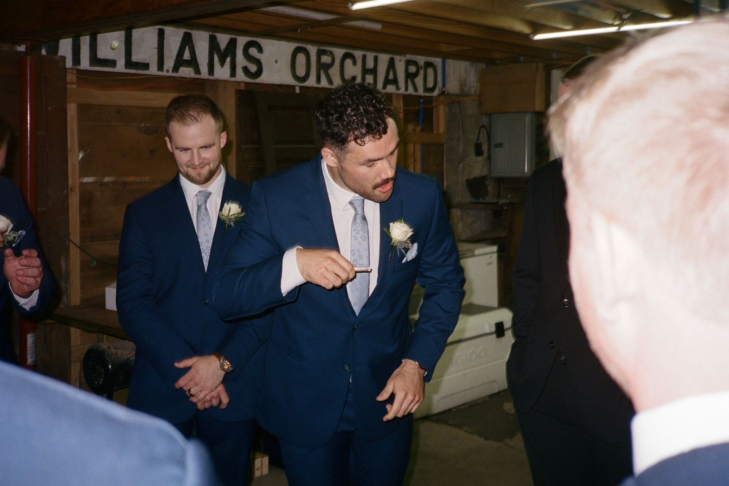 Two groomsmen in navy blue suits with boutonnieres stand inside Williams Orchard barn at a wedding reception.