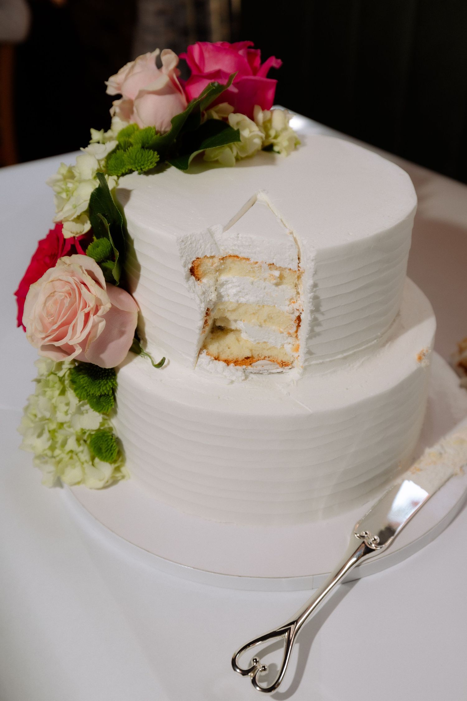 Two-tier white wedding cake with pink roses, slice removed, on white stand with silver cake server.