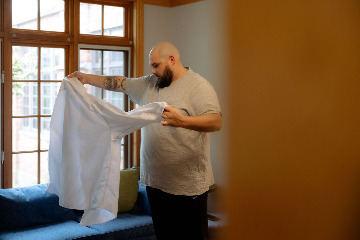 Bald bearded man holding up a white dress shirt near a window with natural light indoors.