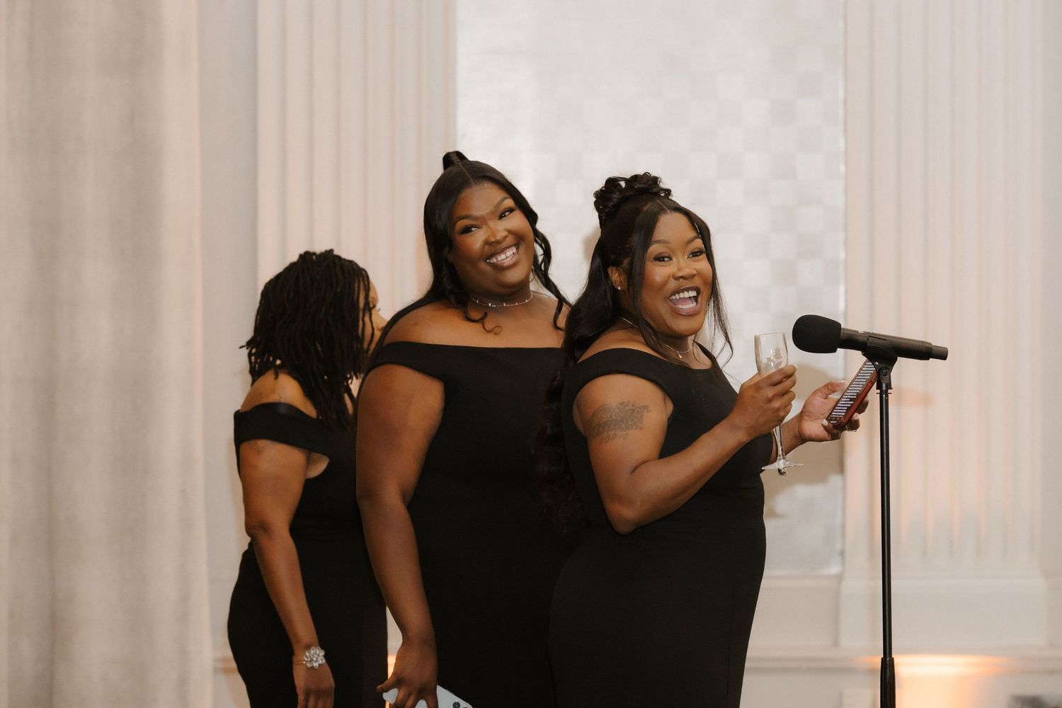 Two women in black off-shoulder dresses smile and speak at a microphone during a formal event.