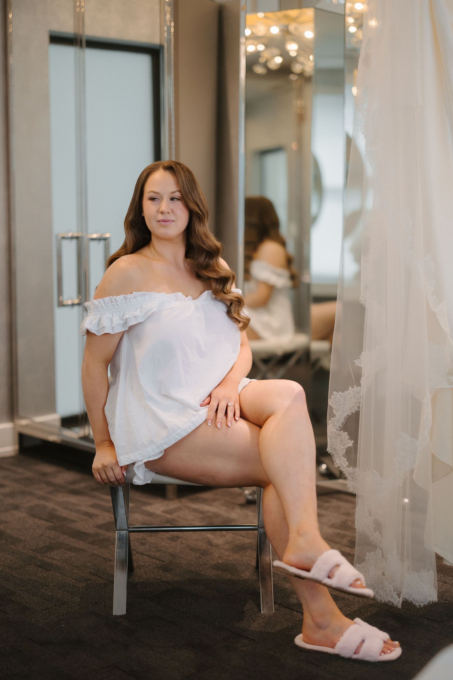 A woman in a white off-shoulder top and pink slippers sits on a chair in a bridal boutique fitting room.