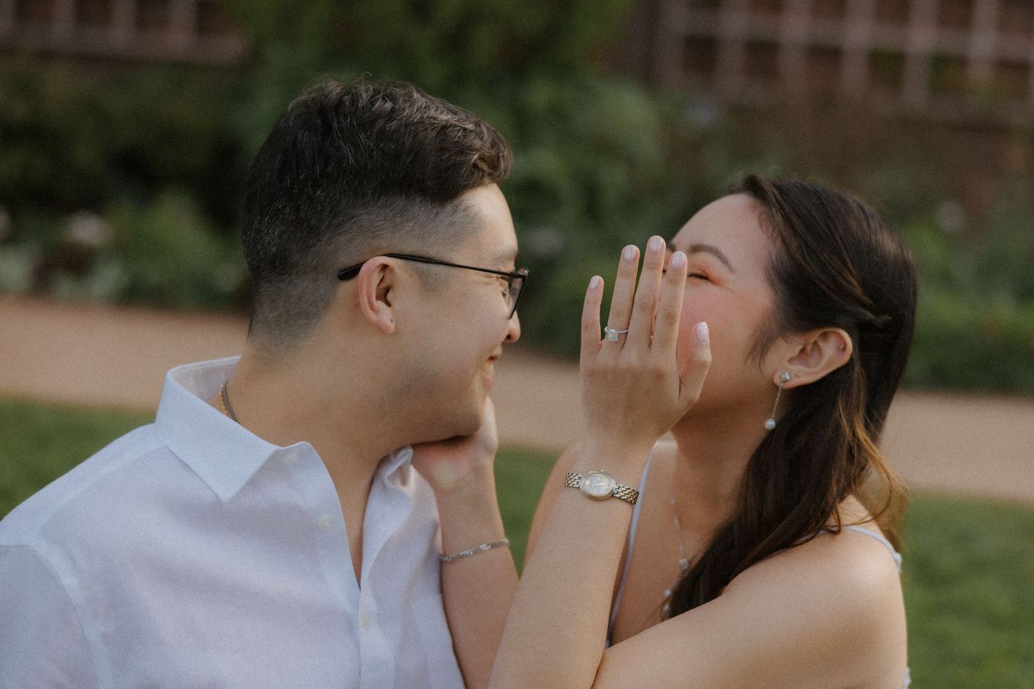A joyful Asian couple sharing a laughing moment outdoors, woman showing off engagement ring while covering her smile.