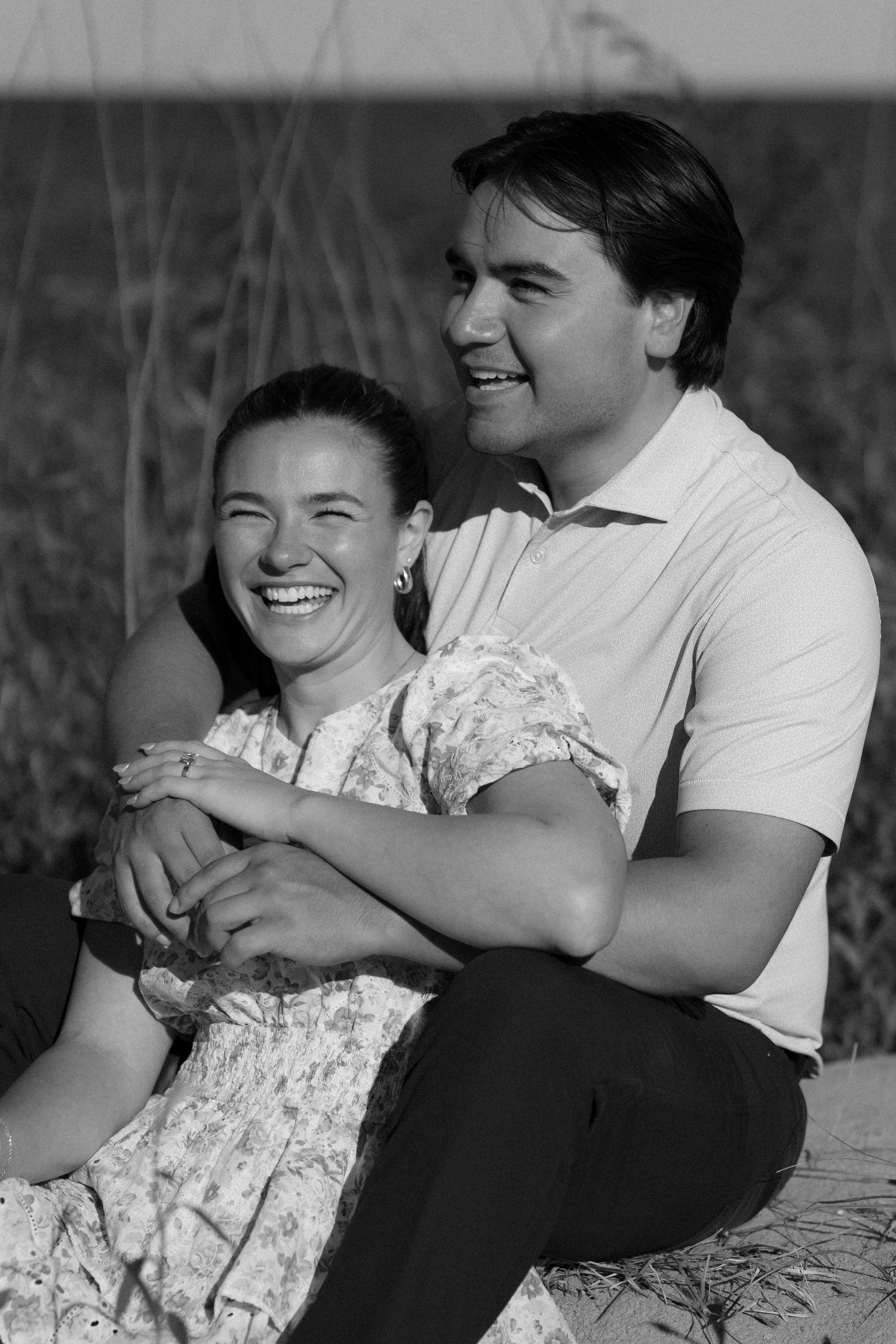 Black and white photo of laughing couple sitting together during golden hour beach session.