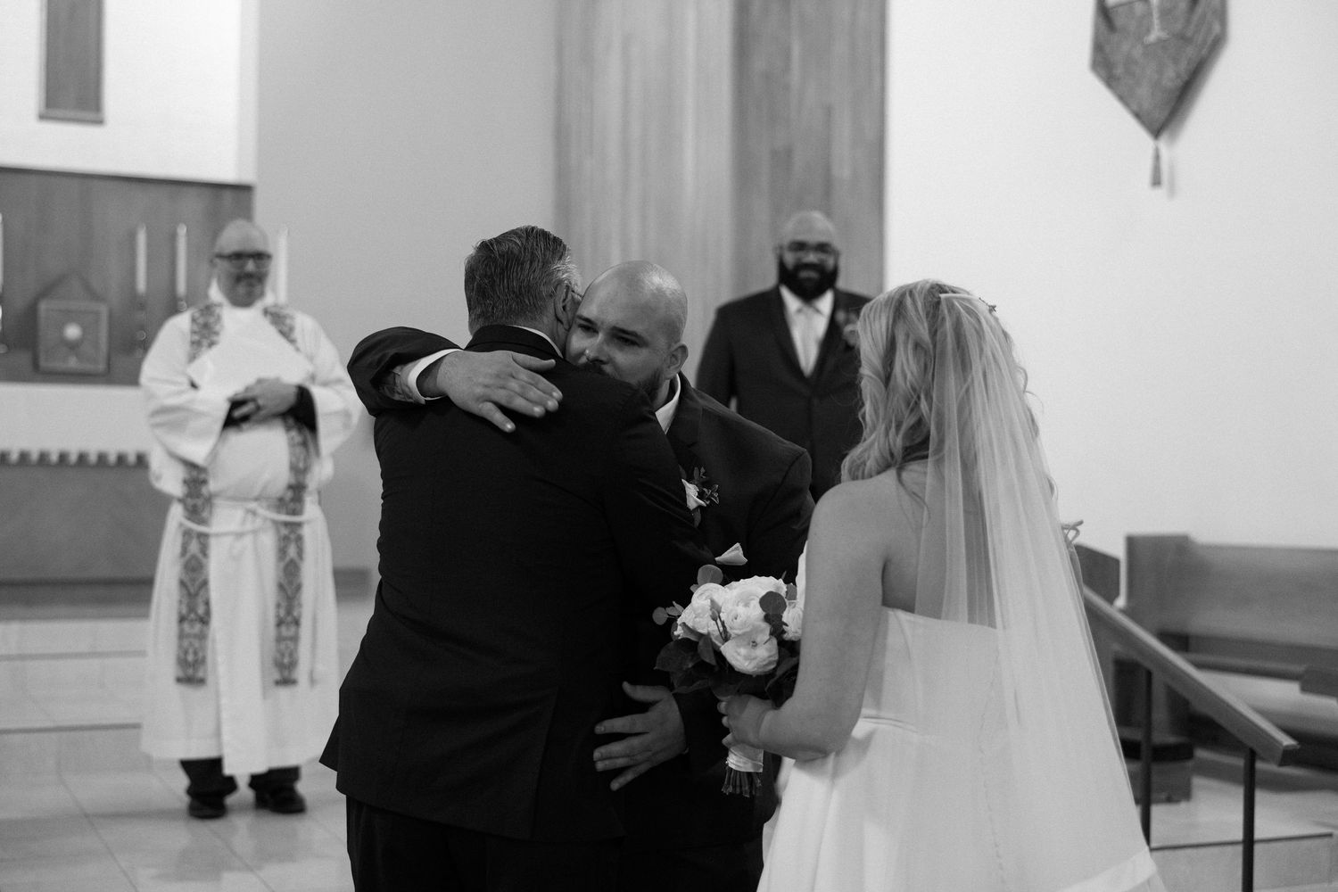 Groom embracing a guest at a church wedding ceremony while bride stands nearby in white gown and veil.