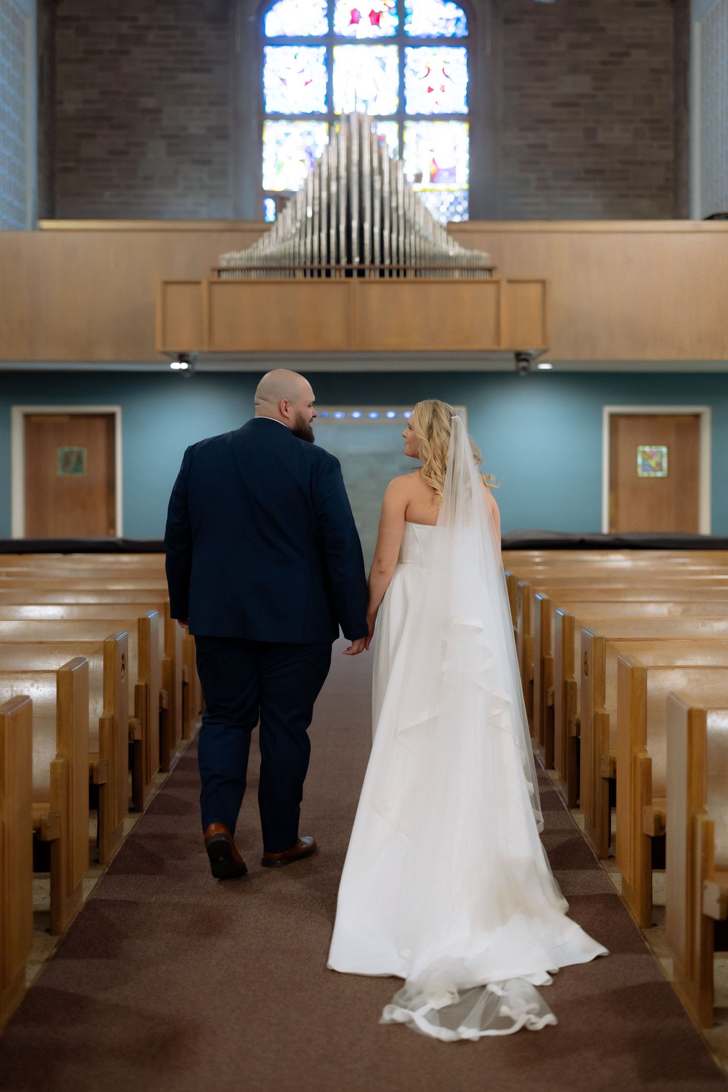 Bride and groom walking hand in hand down church aisle toward pipe organ and stained glass window.