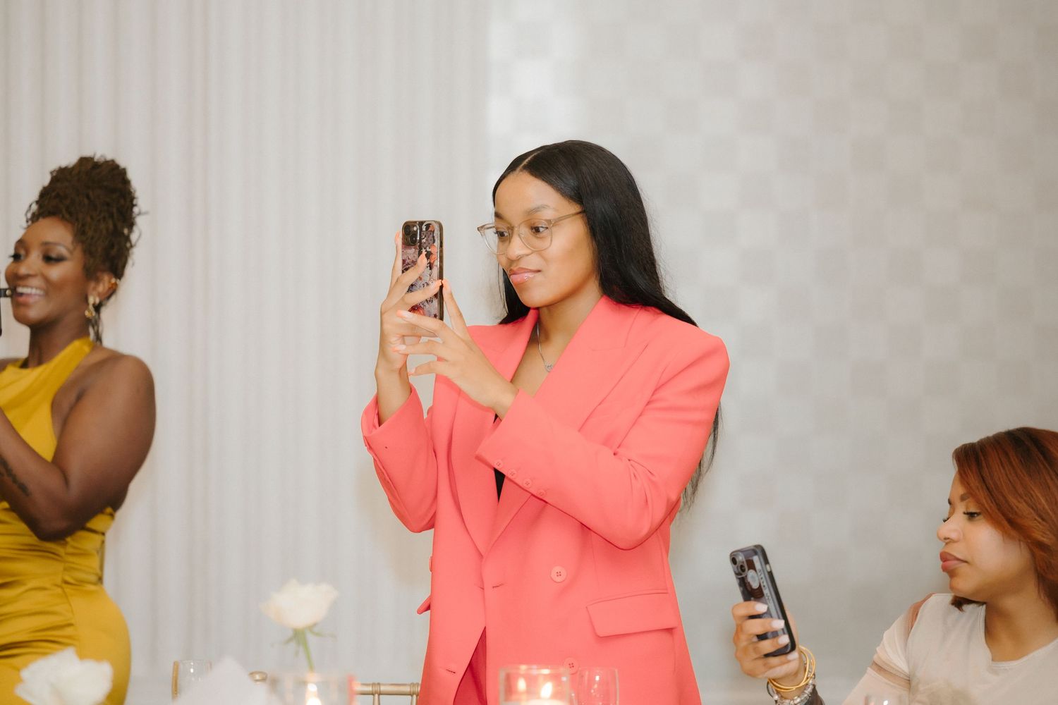 Woman in coral blazer photographing event on smartphone at elegant indoor gathering with guests in background.