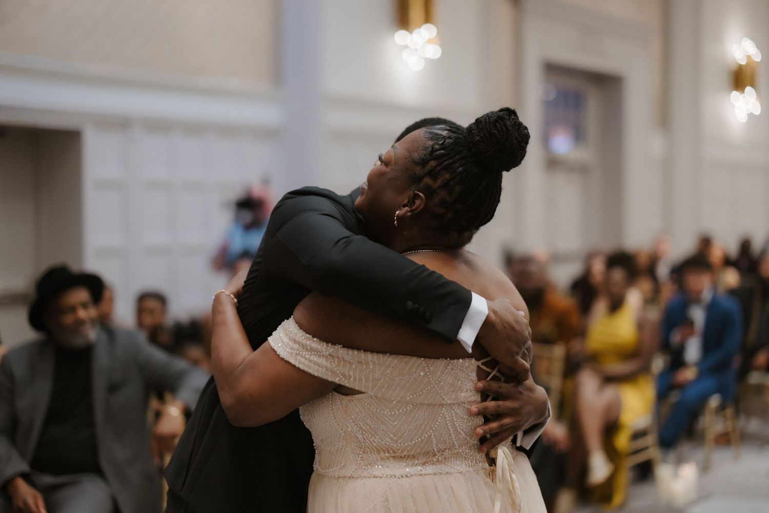 Bride in white lace gown shares emotional hug with guest during wedding ceremony in elegant venue.