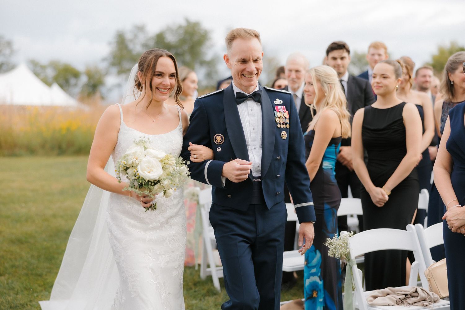 Smiling bride holding white bouquet walks down aisle with military escort in dress uniform past guests.