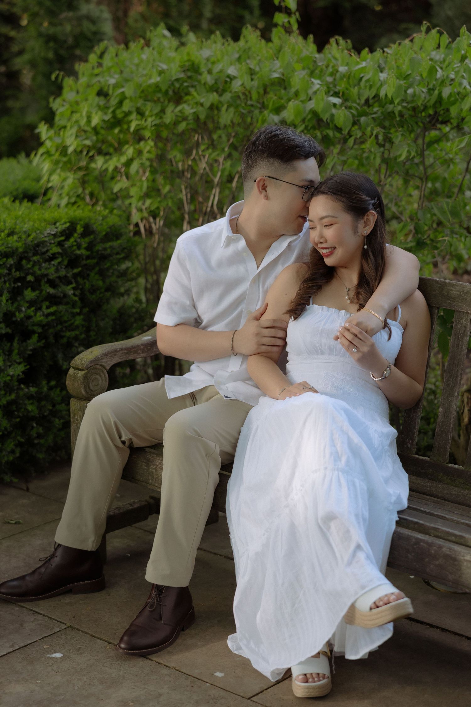 A couple sits on a wooden bench in a garden, man in white shirt kissing woman's cheek as she smiles in a white dress.