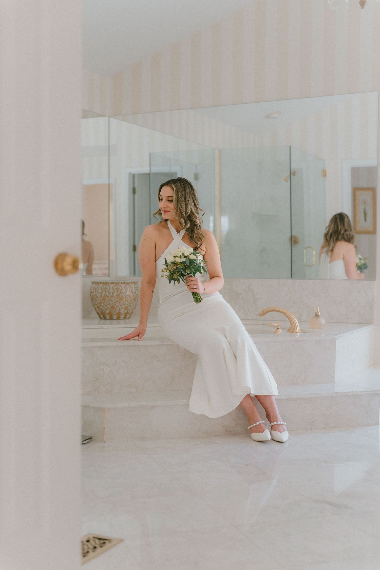 Bride in white gown sitting on marble tub edge holding bouquet in luxury bathroom.