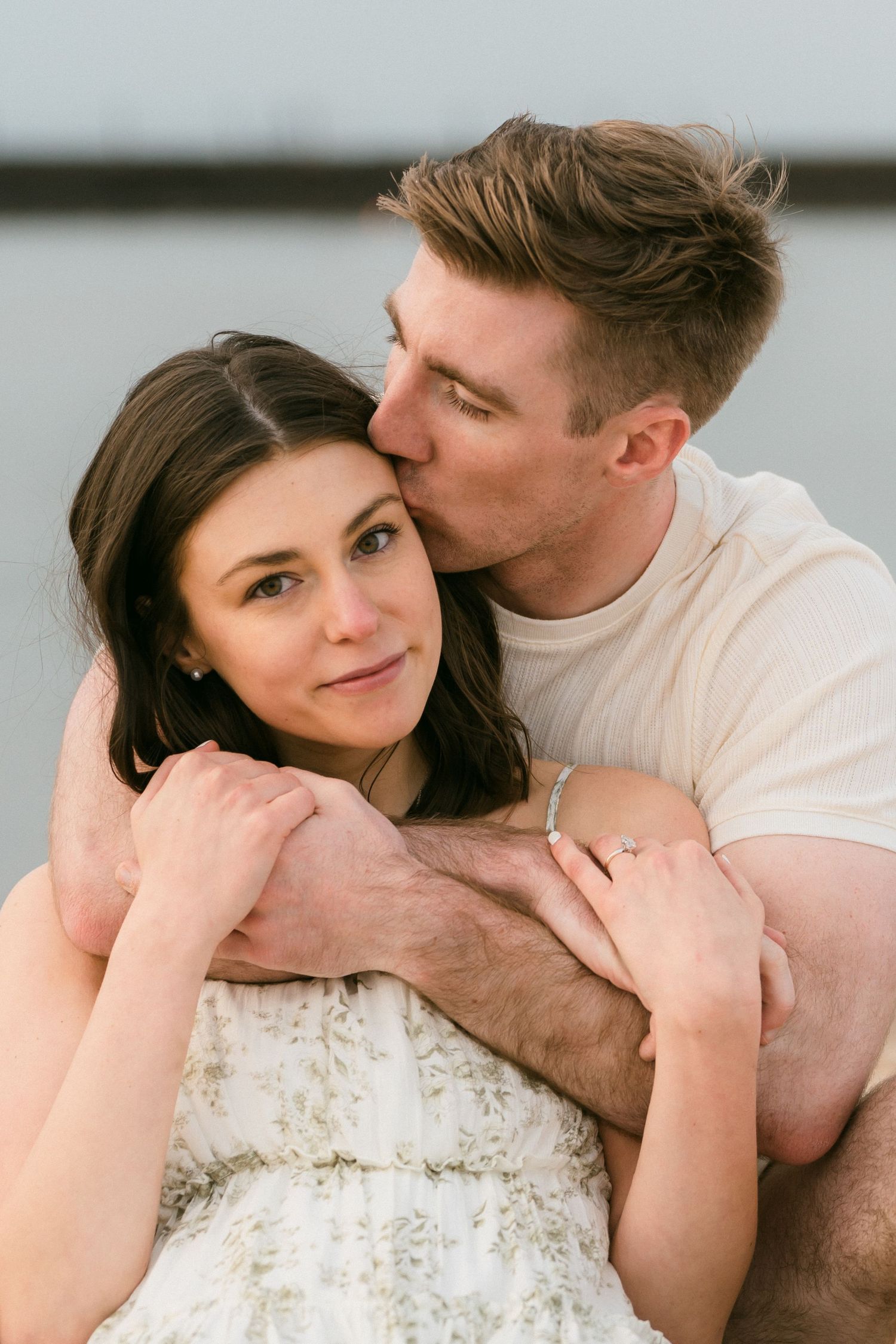 Man tenderly kissing woman on the cheek while embracing her from behind near a serene waterfront setting.