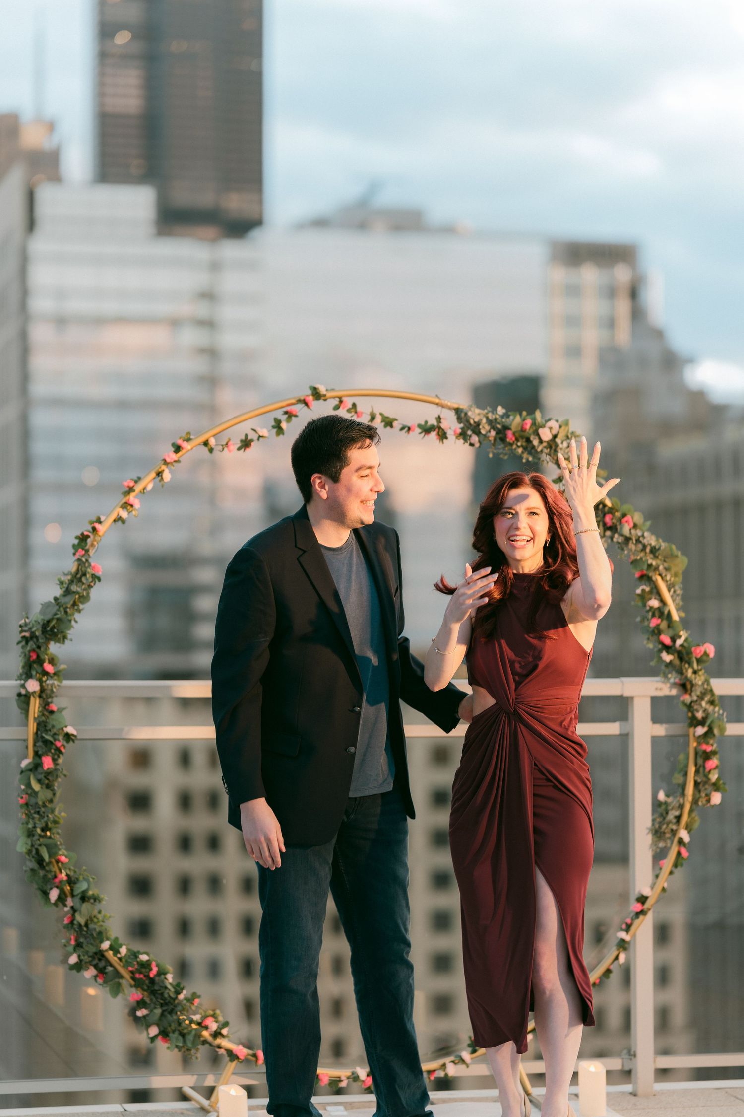 Newly engaged couple smiles as woman shows off engagement ring on Chicago rooftop with floral arch.