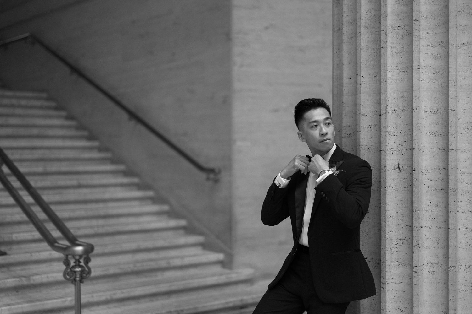 Stylish groom adjusting boutonniere beside grand columns and staircase in black and white photo.