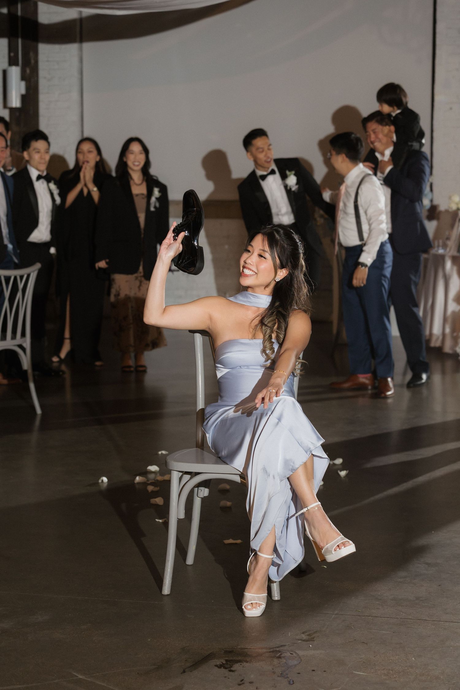 Smiling bridesmaid in blue dress holds up a shoe while seated at wedding reception.