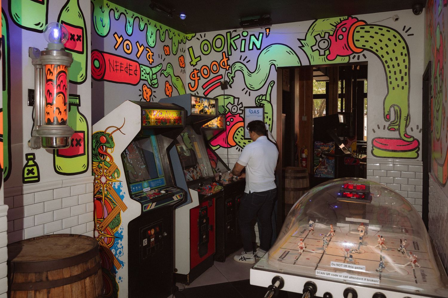 Man playing vintage arcade games in a colorful graffiti-covered retro bar with neon artwork and classic pinball machines.