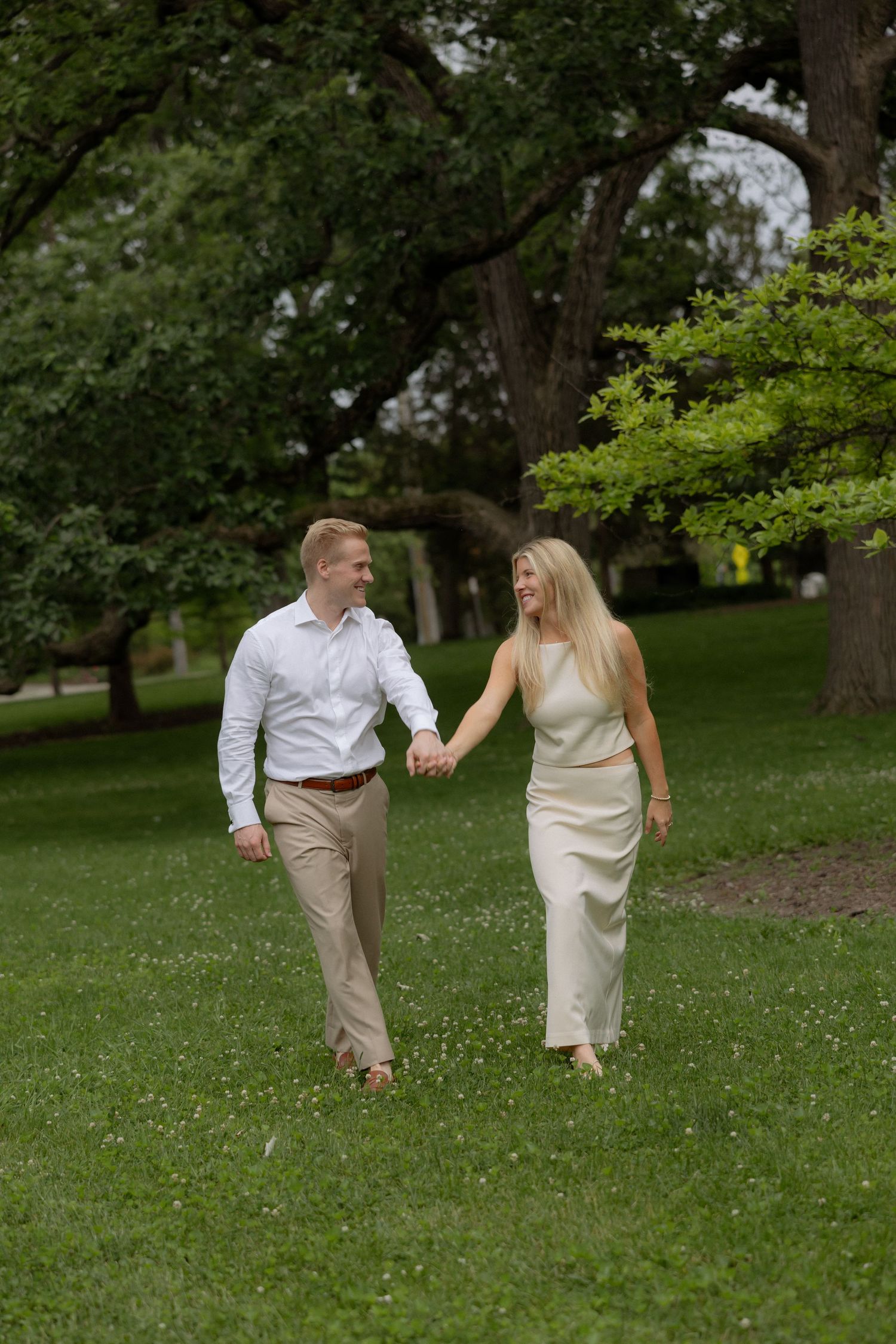 Happy couple holding hands and walking through lush green park, smiling at each other during engagement session.