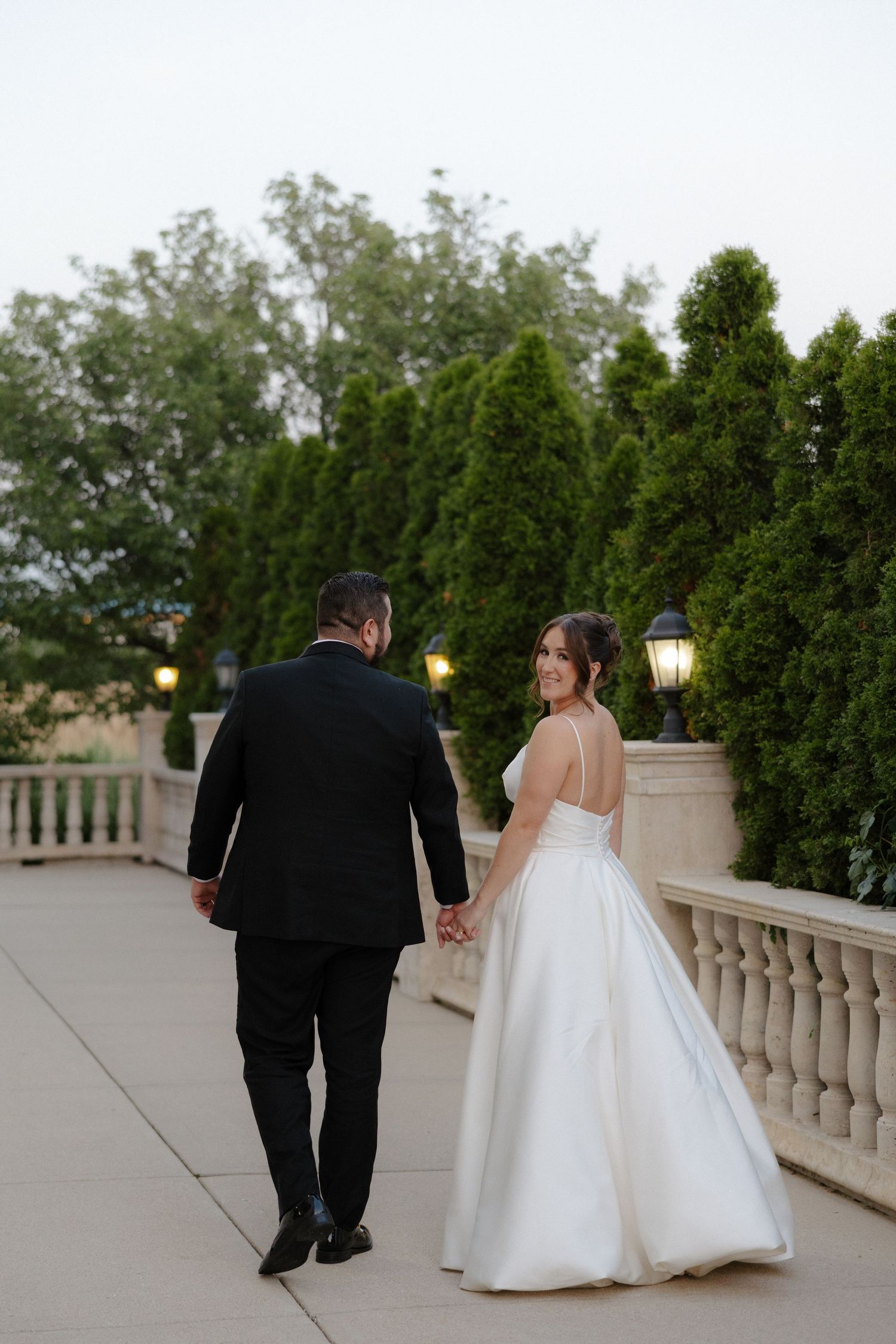 Bride in white ball gown and groom in black suit walk hand in hand on elegant outdoor terrace at dusk.