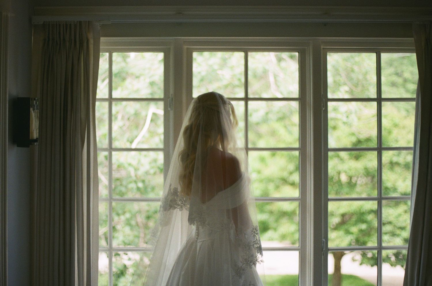 Bride in wedding dress and veil standing at large window overlooking lush garden on her wedding day.