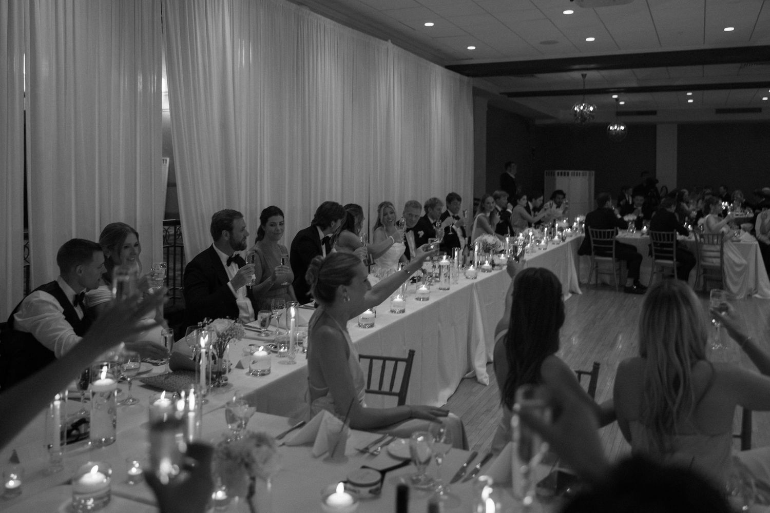 Wedding guests gather at elegantly set reception tables with candles in a softly lit ballroom.