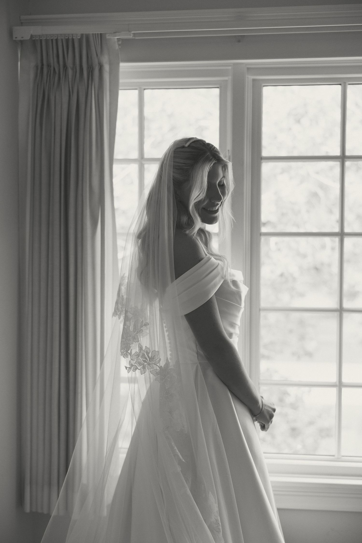 Black and white portrait of a smiling bride in veil and off-shoulder gown standing by a window.