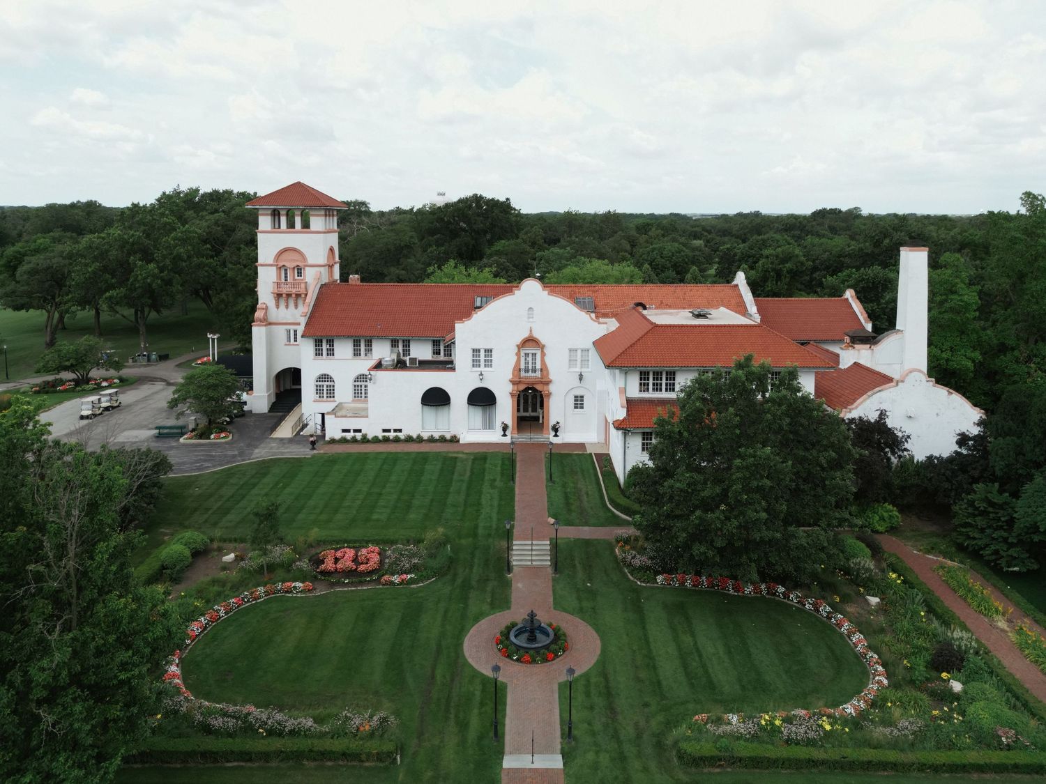 Aerial view of a grand Spanish Colonial Revival mansion with red tile roof, manicured gardens, and brick pathways on lush grounds.