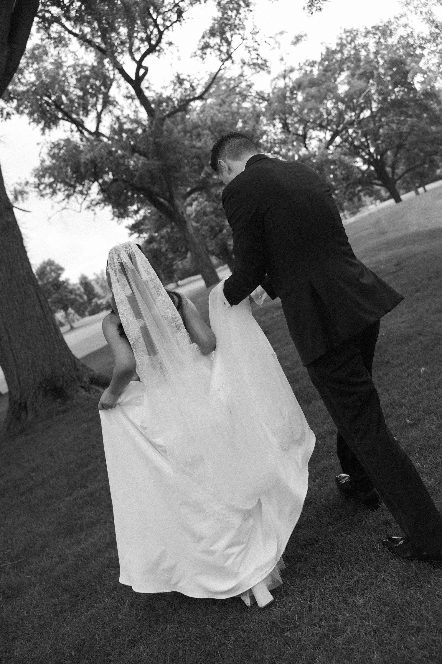 Groom assists bride with wedding dress train while walking through park in black and white photo.