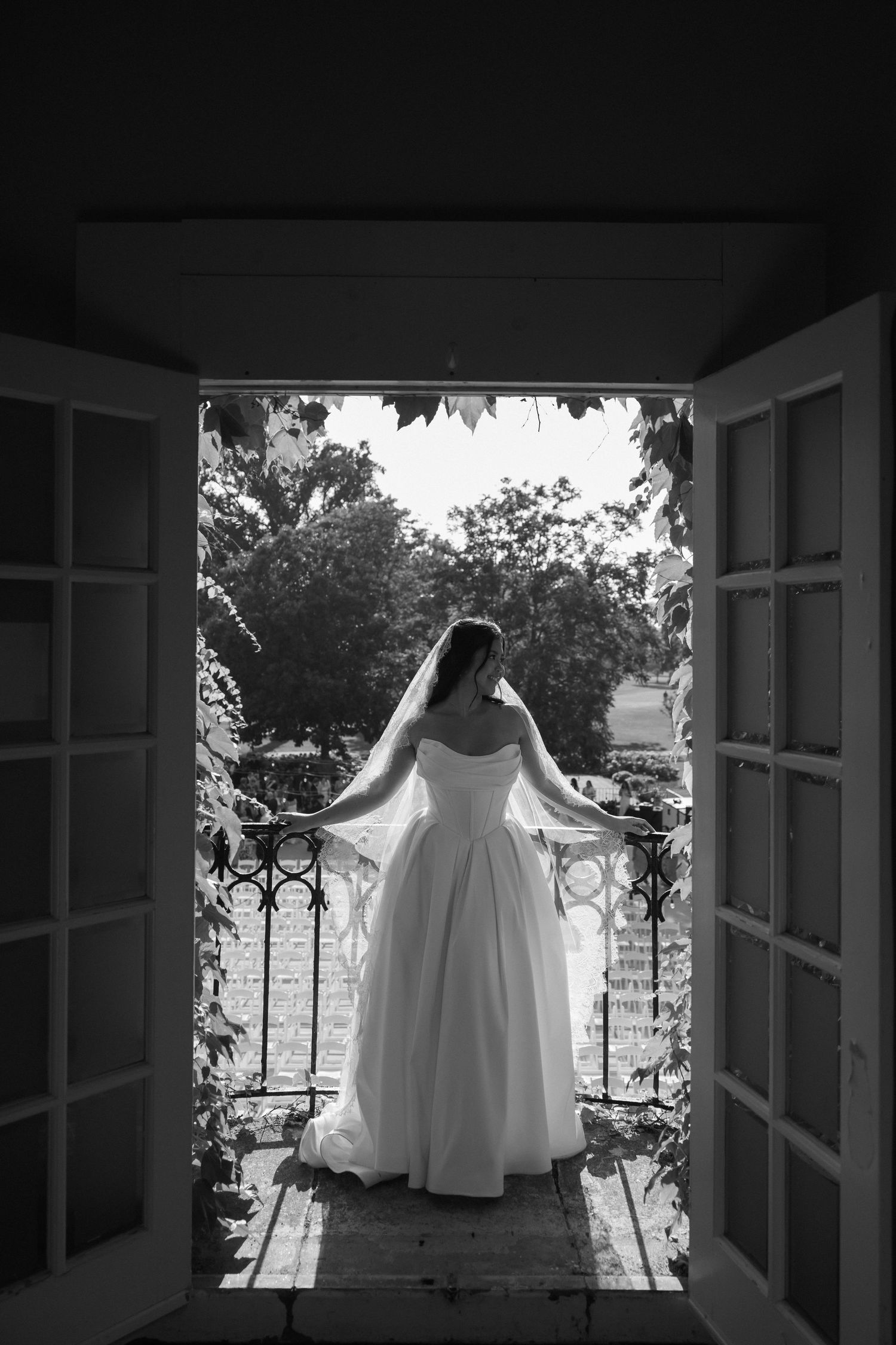 Bride in white gown and veil stands on balcony with ornate railing, black and white wedding photo.
