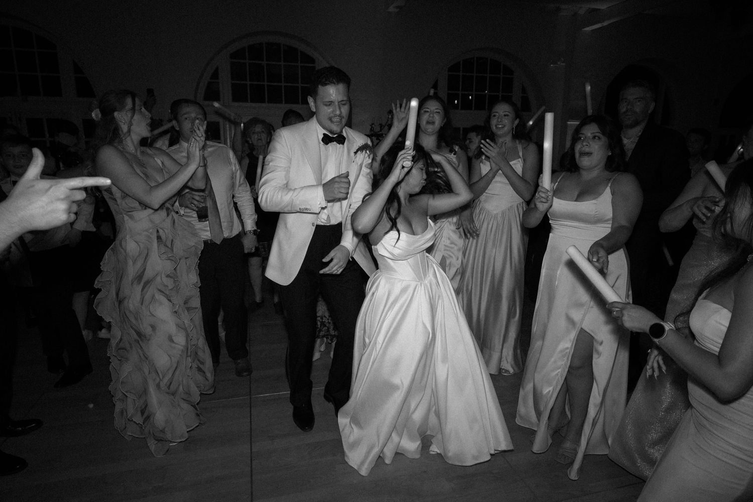 Black and white photo of wedding party dancing with candles during lively indoor reception celebration.