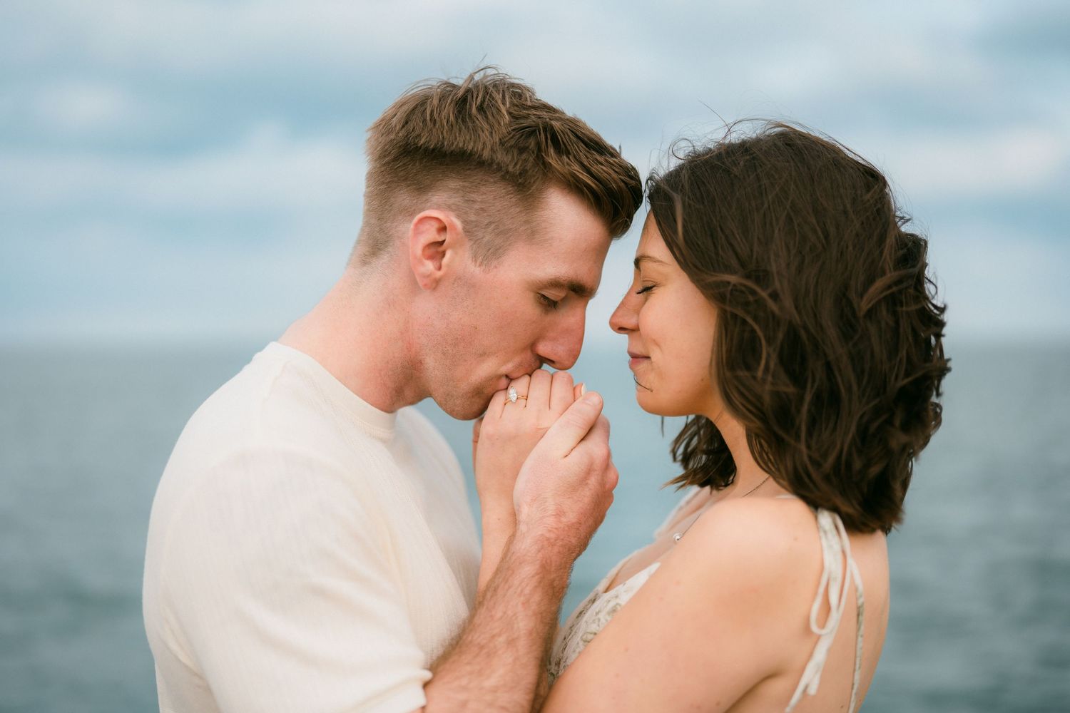 Man gently kissing woman's hand near a calm blue lake outdoors.