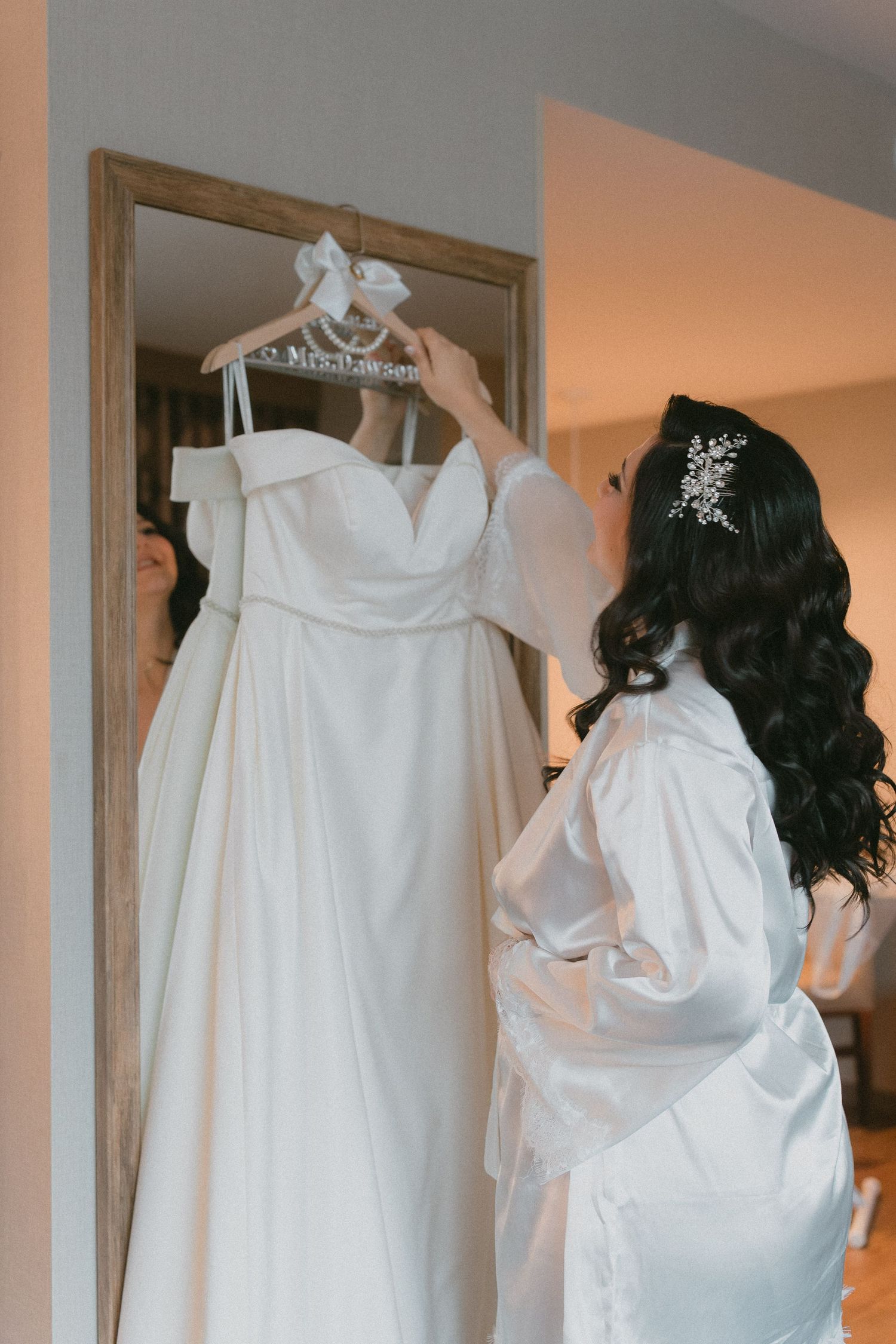 Bride in white robe admires her off-shoulder wedding gown hanging on a personalized hanger in a mirror.