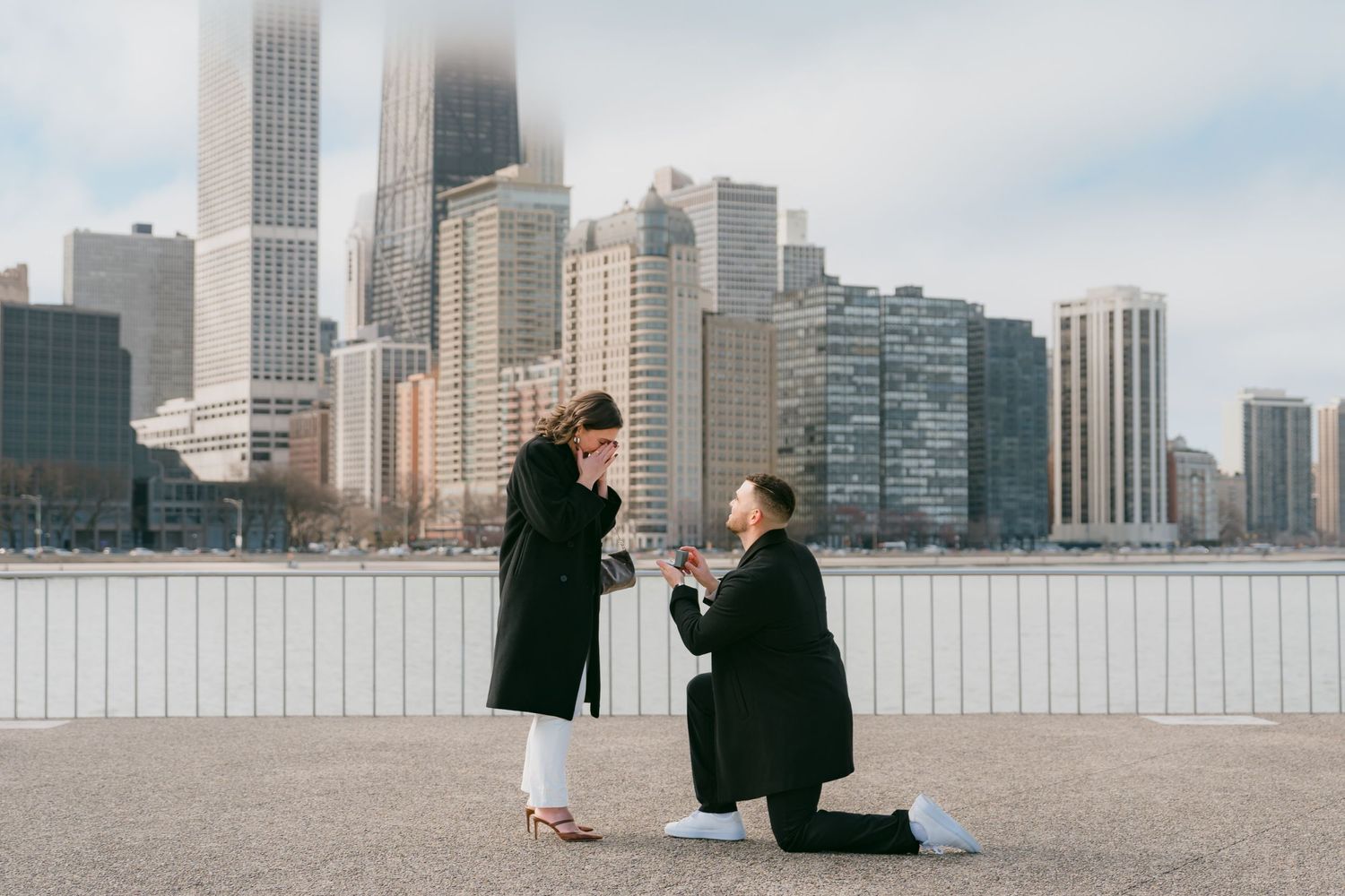 Man proposing on one knee to surprised woman on Chicago lakefront with city skyline backdrop.