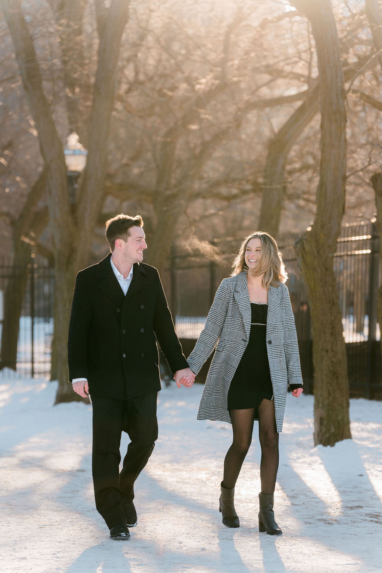 Happy couple holding hands and walking through a sunlit snowy park path in winter.