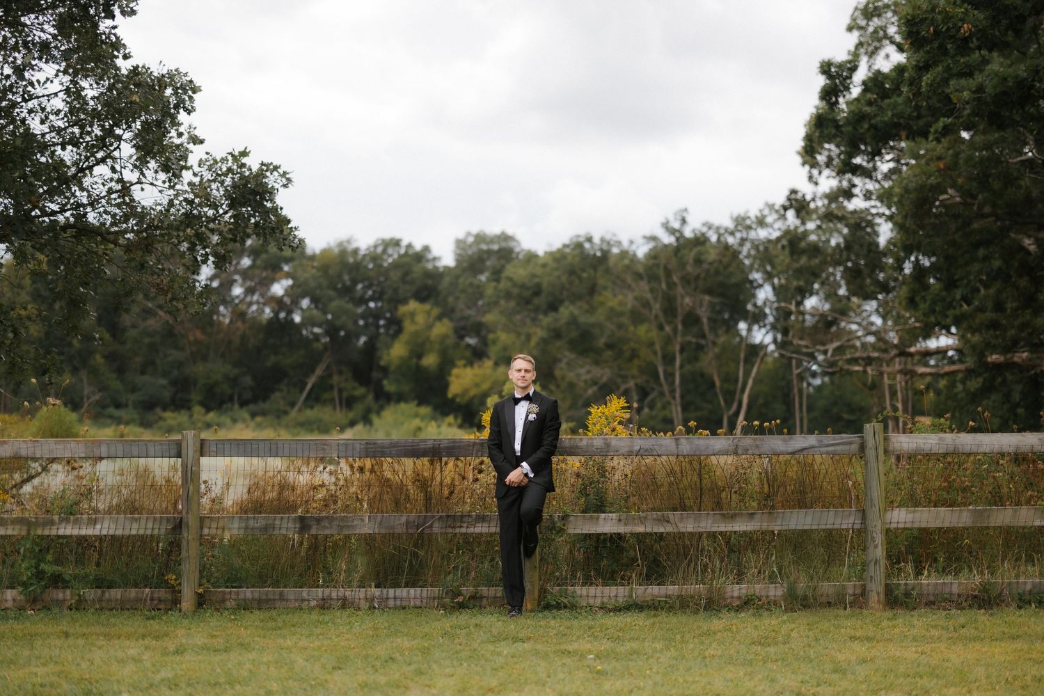 Groom in black tuxedo standing alone by wooden fence in expansive green field on wedding day.