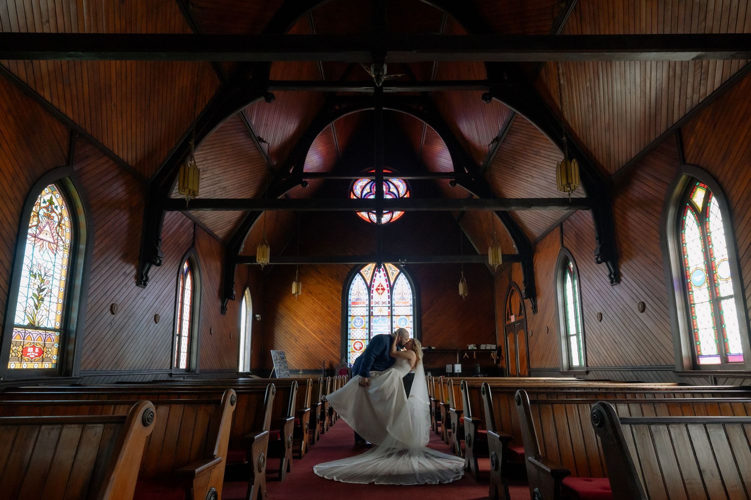 A couple shares a romantic dip kiss in a stunning wooden church with stained glass windows during their wedding ceremony.