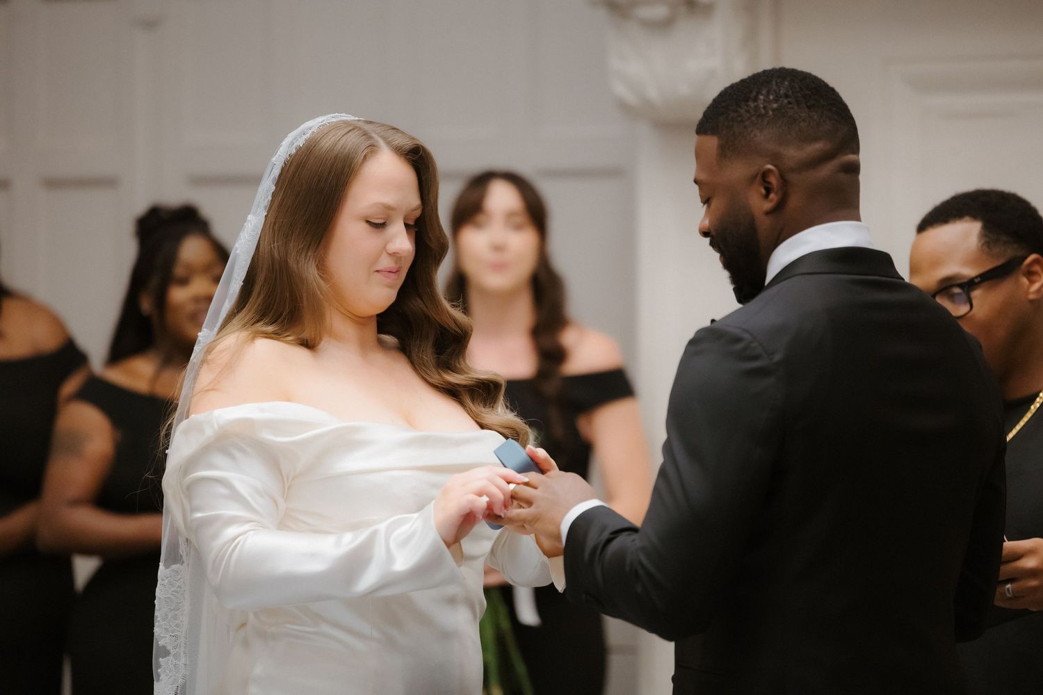 Groom placing wedding ring on bride's finger during ceremony, bride in white gown and veil, guests in black attire.