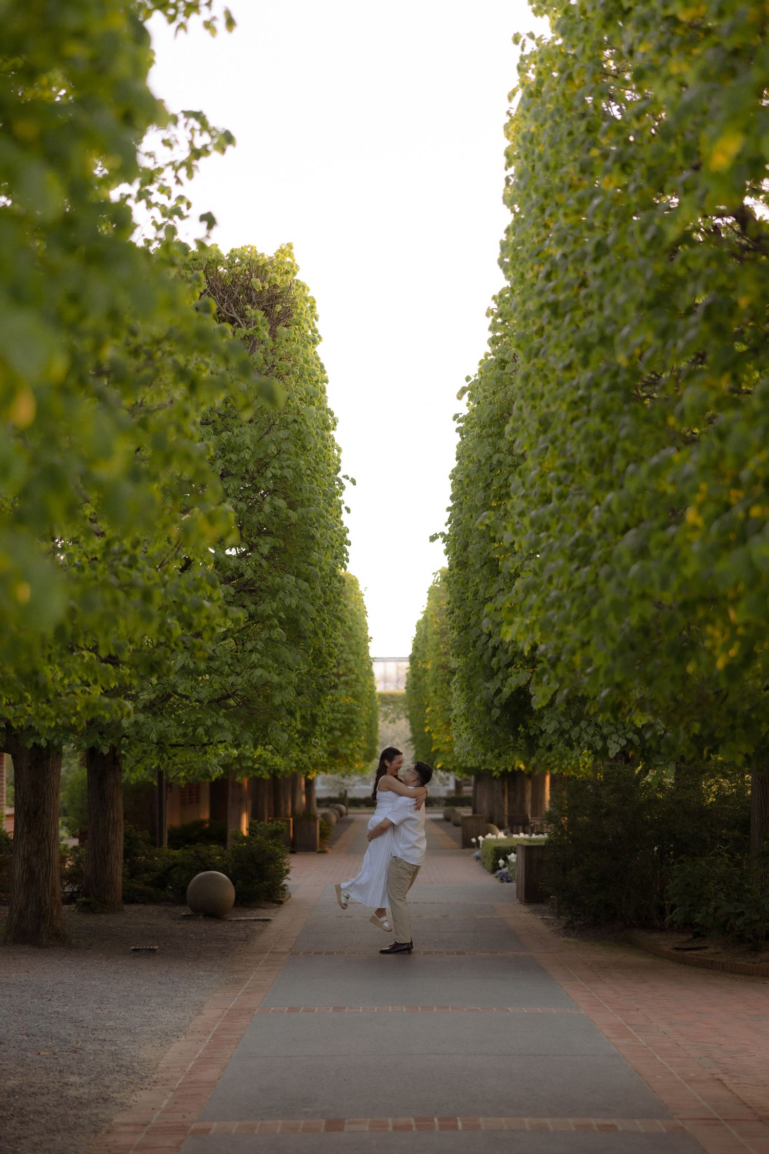 Couple dancing on a tree-lined garden path, surrounded by lush green trees in a romantic outdoor setting.